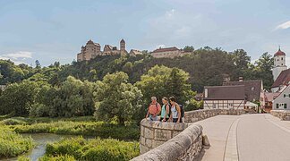 Drei Wanderer stehen auf der steinernen Brücke und blicken auf die Wörnitz. Links oben thront über dem Wald die Harburg.