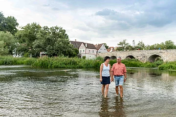 Ein Paar watet am Wörnitzstrand durchs Wasser. Im Hintergrund sieht man die steinerne Brücke und Häuser von Harburg.
