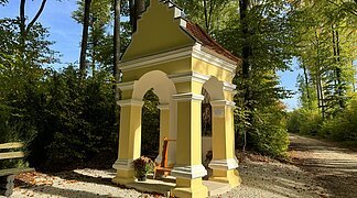 Eine gelbe kleine Kapelle steht an einem Waldweg. Außemherum sind grün belaubte Bäume.