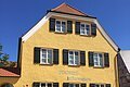 Blick auf das gelbe Gebäude der Tourist-Information Wemding. Im Vordergrund plätschert ein Brunnen und stehen Bänke.