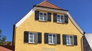 Blick auf das gelbe Gebäude der Tourist-Information Wemding. Im Vordergrund plätschert ein Brunnen und stehen Bänke.