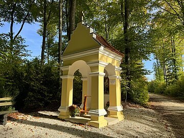 Waldkapelle Wolferstadt-Wemding Eine gelbe kleine Kapelle steht an einem Waldweg. Außemherum sind grün belaubte Bäume.