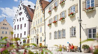 Zwei Freundinnen sitzen auf einer Bank vor dem Verwaltungsgebäude am historischen Marktplatz und unterhalten sich.