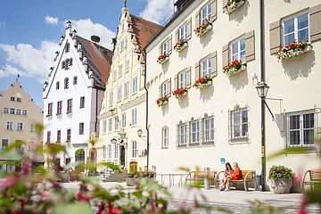 Zwei Freundinnen sitzen auf einer Bank vor dem Verwaltungsgebäude am historischen Marktplatz und unterhalten sich.