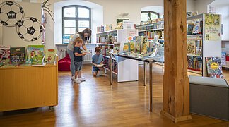 Eine Frau ist mit ihren beiden Kindern in der Stadtbücherei. Sie suchen sich Bücher zur Ausleihe heraus.