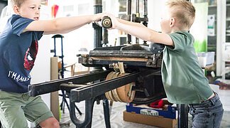 Zwei Jungs testen den Bilddruck mit einer händischen Maschine im KunstMuseum Donau-Ries.