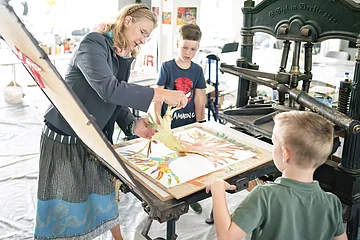 Die Leiterin des KunstMuseums führt zwei Jungs den Bilddruck mit einer alten Maschine vor.