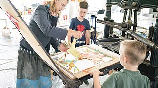 Die Leiterin des KunstMuseums führt zwei Jungs den Bilddruck mit einer alten Maschine vor.