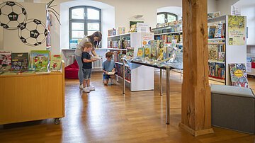 Stadtbücherei Eine Frau ist mit ihren beiden Kindern in der Stadtbücherei. Sie suchen sich Bücher zur Ausleihe heraus.