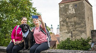 Zwei Freundinnen sitzen mit ihrem Hund auf der Stadtmauer und machen ein Selfie mit dem Baronturm.