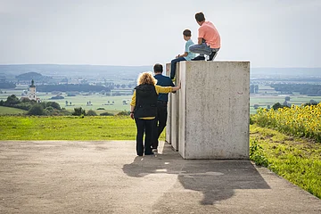 Die Kinder sitzen auf den Steinen der Zeitpyramide und die Eltern stehen daneben. Gemeinsam blicken sie in das Ries und genießen den tollen Ausblick.