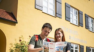 Zwei Touristen stehen vor der Tourist-Information in Wemding und schauen sich die Imagebroschüre gemeinsam an.