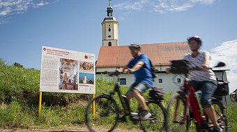 Zwei Radfahrer fahren an der Informationstafel des Sagenwegs vorbei.