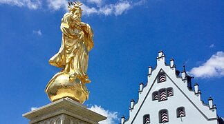 Marktplatz Wemding Hoch aufragende Fassade des historischen Rathaus mit einem blauen Himmel im Hintergrund. Im Vordergrund steht eine goldene Marienstatue mit Brunnen.