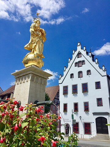 Hoch aufragende Fassade des historischen Rathaus mit einem blauen Himmel im Hintergrund. Im Vordergrund steht eine goldene Marienstatue mit Brunnen.