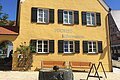 Blick auf das gelbe Gebäude der Tourist-Information Wemding. Im Vordergrund plätschert ein Brunnen und stehen Bänke.