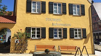 Blick auf das gelbe Gebäude der Tourist-Information Wemding. Im Vordergrund plätschert ein Brunnen und stehen Bänke.