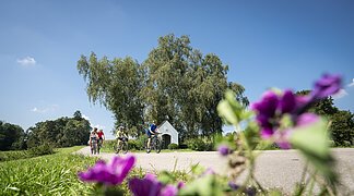Kapelle Warching, Monheim Drei Personen fahren auf Fahrrädern auf einem Weg, im Hintergrund Bäume und eine kleine Kapelle. Im Vordergrund unscharfe Blumen.