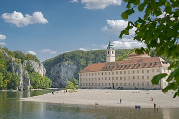 Flussufer mit Kiesstrand, altes Gebäude mit Turm und bewaldete Felsen unter blauem Himmel mit Wolken