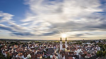 Panoramablick auf Stadt mit roten Dächern und zwei Kirchtürmen bei Sonnenuntergang unter bewölktem Himmel