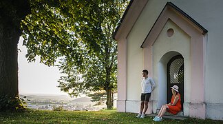 Wanderer auf der Monheimer Alb Mann lehnt an Hauswand, Frau sitzt in Türbogen, große Bäume, Dorf im Hintergrund bei Tageslicht.