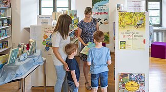 Stadtbüchereileitung Frau Hertle berät eine Frau mit ihren Kindern zu bestimmten Büchern.
