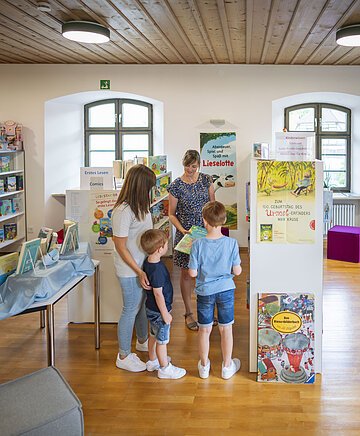 Stadtbüchereileitung Frau Hertle berät eine Frau mit ihren Kindern zu bestimmten Büchern.