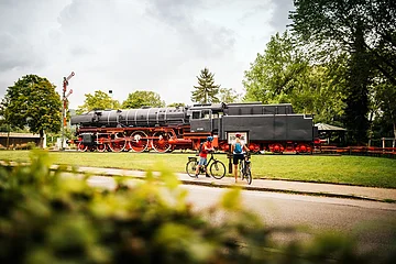 Dampflokomotive auf Gleis mit zwei Radfahrern davor, umgeben von Bäumen und Wiese bei bewölktem Himmel.