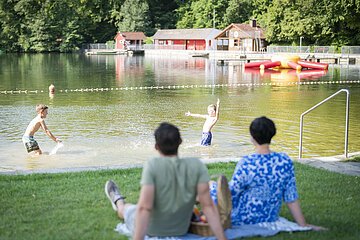 Ein Ehepaar sitzt auf der Liegewiese am Waldsee und beobachtet ihre spielenden Kinder im Wasser am Waldsee.