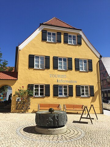 Blick auf das gelbe Gebäude der Tourist-Information Wemding. Im Vordergrund plätschert ein Brunnen und stehen Bänke.